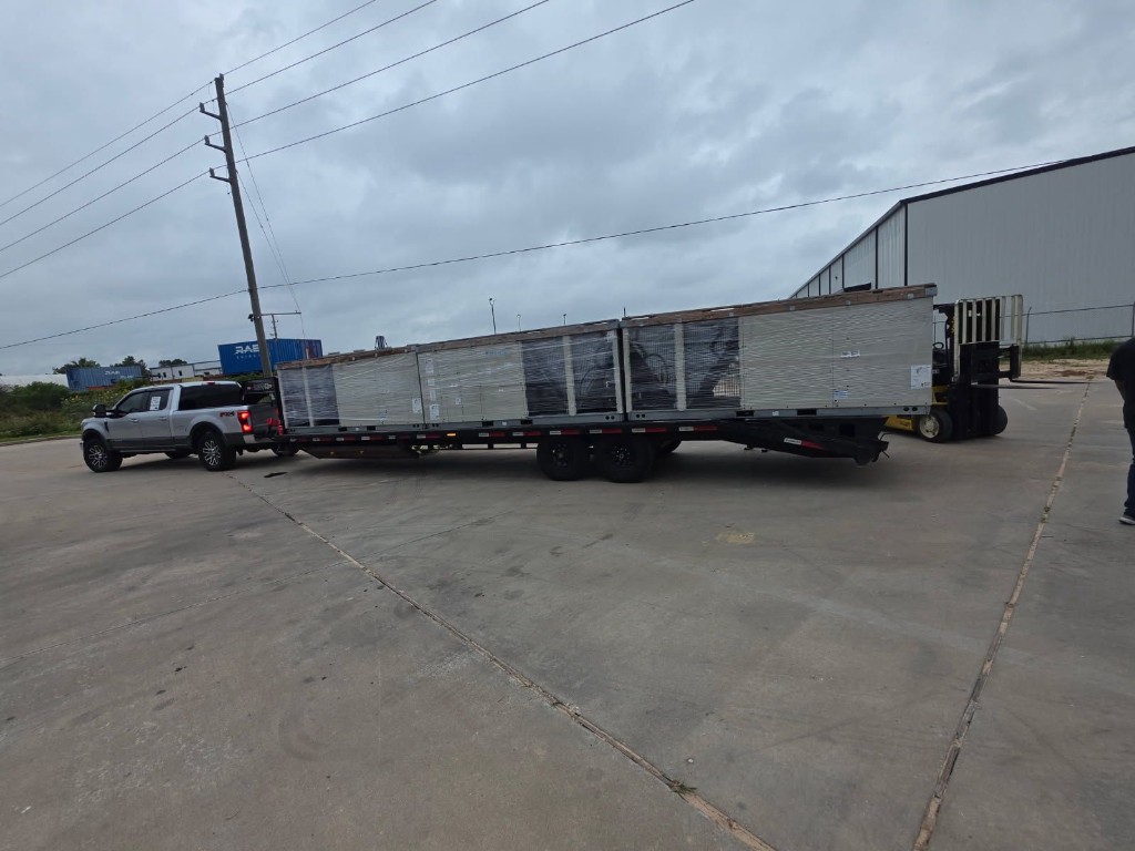 Side view of flatbed trailer loaded with wrapped industrial units at a paved commercial facility.