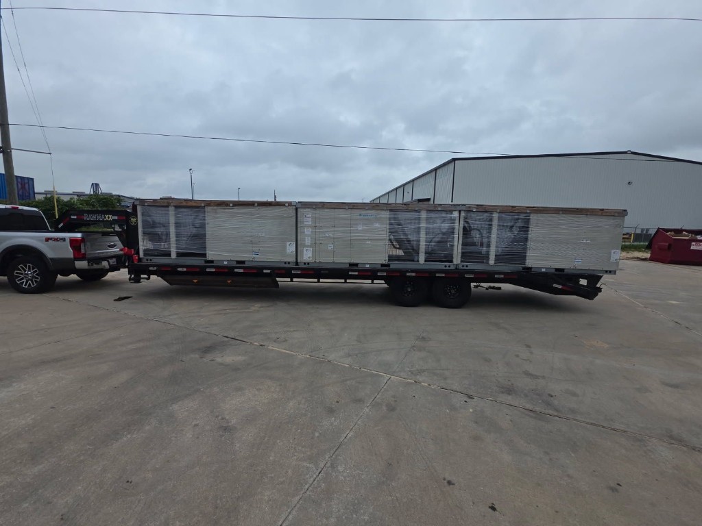 Gooseneck flatbed trailer loaded with shrink-wrapped commercial HVAC-style equipment behind a pickup truck.