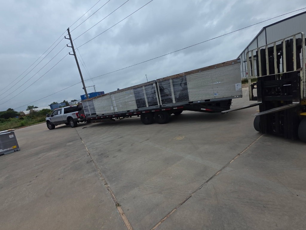 Wide view of pickup and gooseneck trailer with multiple large wrapped units and warehouse in the background.