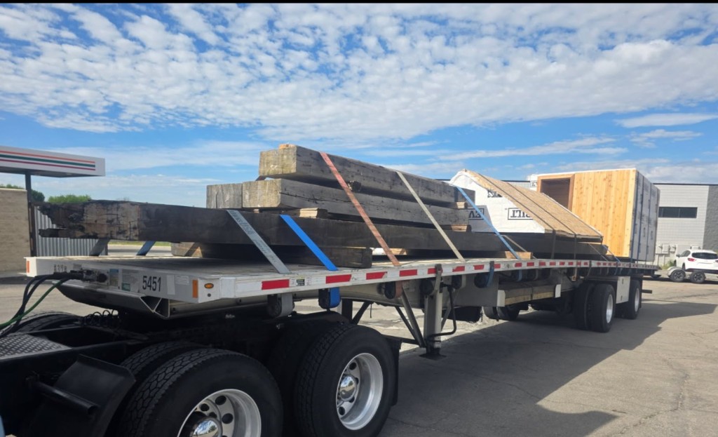 Side view of flatbed trailer with heavy timber beams and modular unit secured with colored straps.