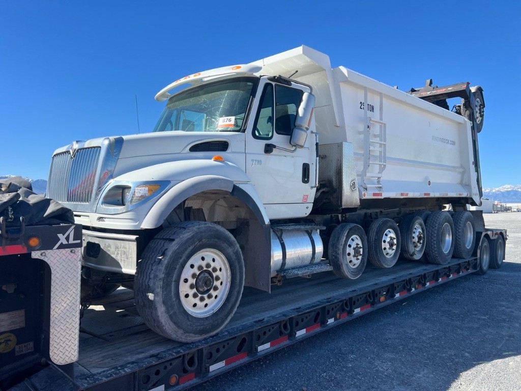 Front three-quarter view of a white heavy-duty dump truck loaded on a black multi-axle lowboy trailer under a clear sky.