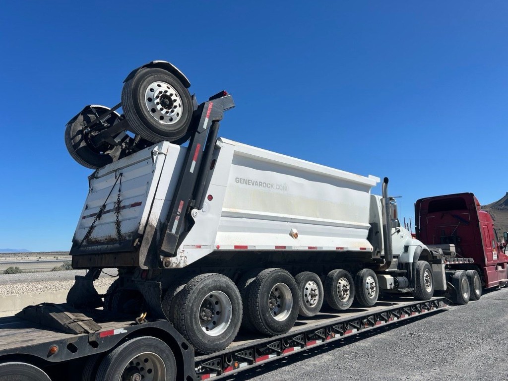 Side view of dump truck on lowboy with raised auxiliary axle assembly visible on the load.