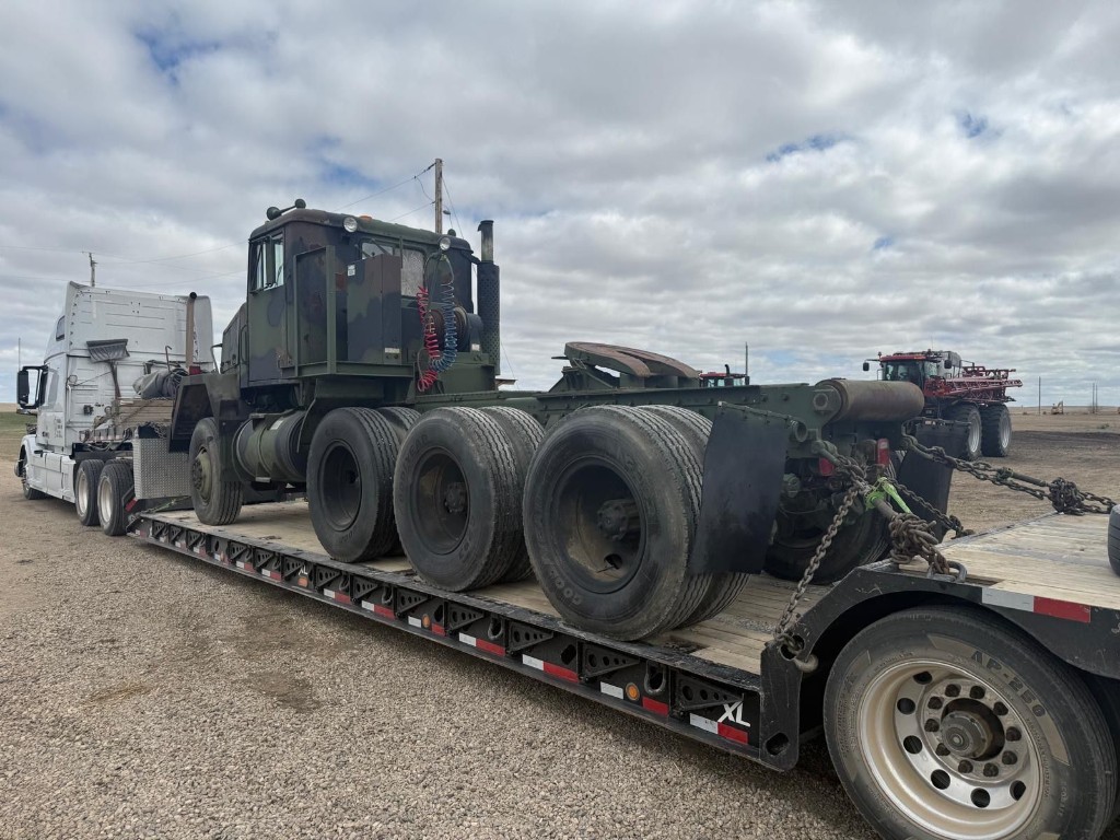 Rear three-quarter view of camouflage military truck on lowboy with chain securement visible.
