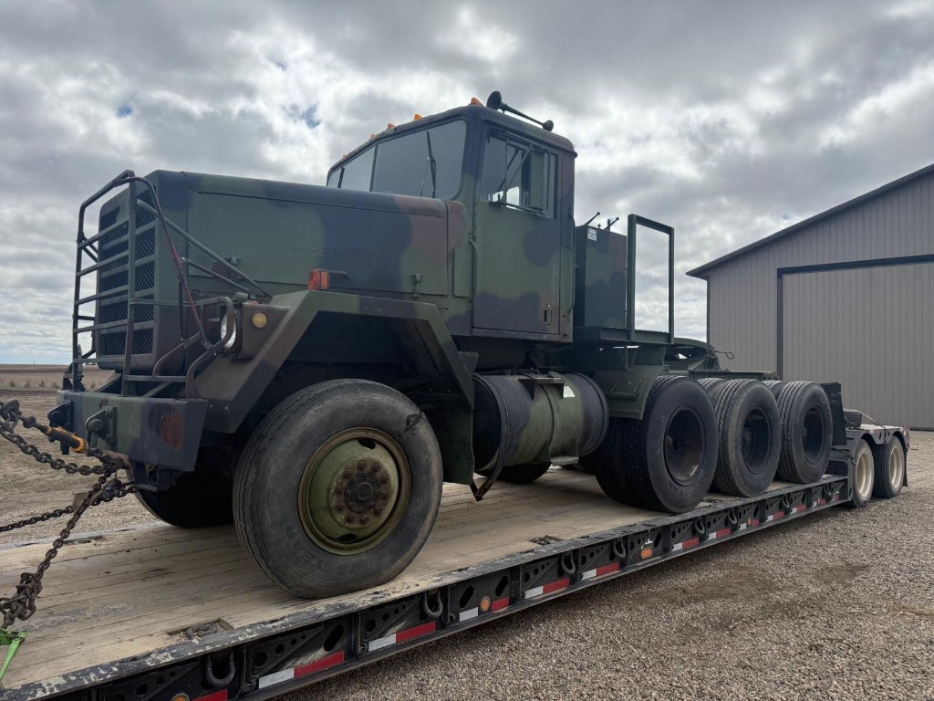 Military-style heavy truck chained to lowboy in front of industrial building.
