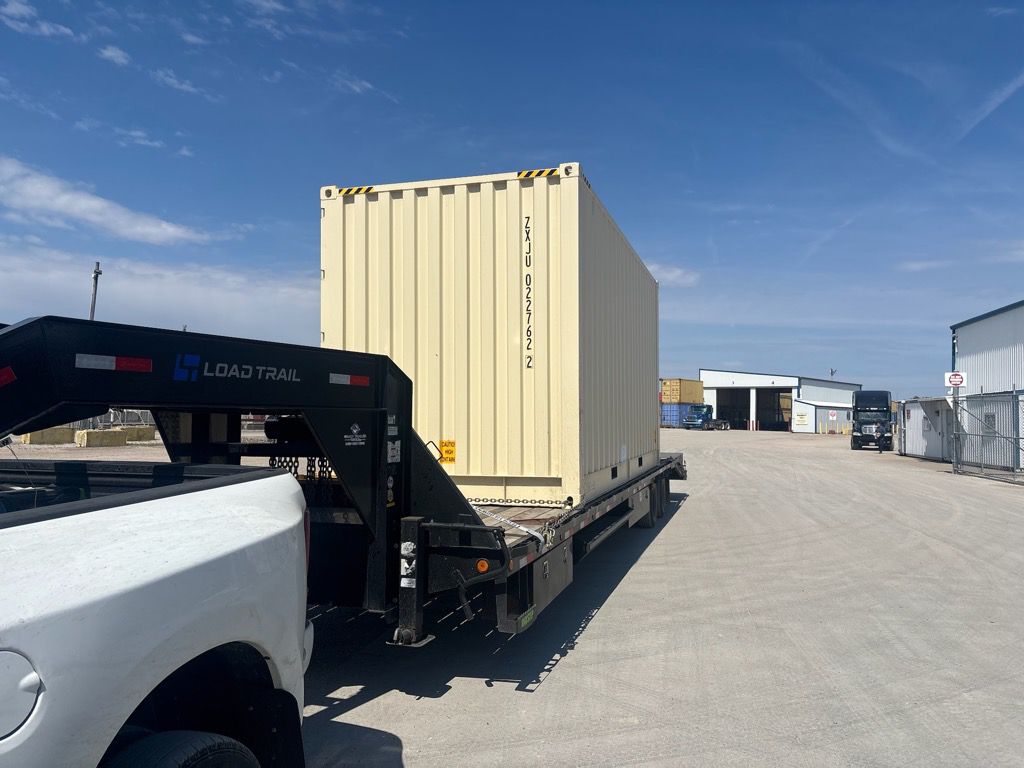 Shipping container secured on a gooseneck flatbed trailer behind a pickup truck at a commercial lot.